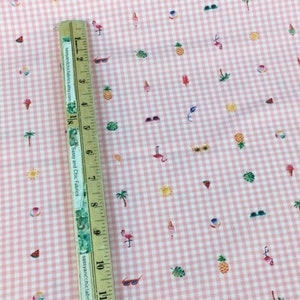 Puede incluir: Tela de cuadros rosa y blanco con un estampado de verano. El dise&ntilde;o incluye peque&ntilde;as ilustraciones de gafas de sol, pi&ntilde;as, helados, flamencos y palmeras. Una regla de madera se coloca en diagonal sobre la tela, midiendo hasta 28 cm.