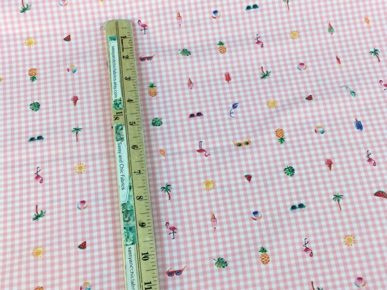 Puede incluir: Tela de cuadros rosa y blanco con un estampado de verano. El dise&ntilde;o incluye peque&ntilde;as ilustraciones de gafas de sol, pi&ntilde;as, helados, flamencos y palmeras. Una regla de madera se coloca en diagonal sobre la tela, midiendo hasta 28 cm.