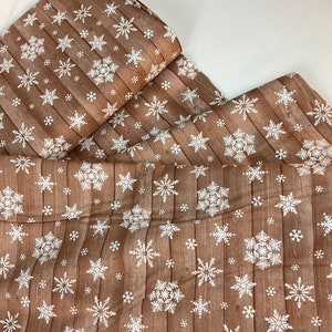 Pu&ograve; includere: Tessuto marrone con motivo a venature di legno e fiocchi di neve bianchi. Il tessuto &egrave; piegato, mostrando il motivo in dettaglio. I fiocchi di neve variano per dimensioni e design, creando un tema invernale o festivo.