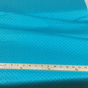 May include: Turquoise fabric with a repeating pattern of small, darker blue dots. A wooden ruler with inch markings is placed across the fabric. The ruler has text that reads "Sassy and Chic Fabrics".