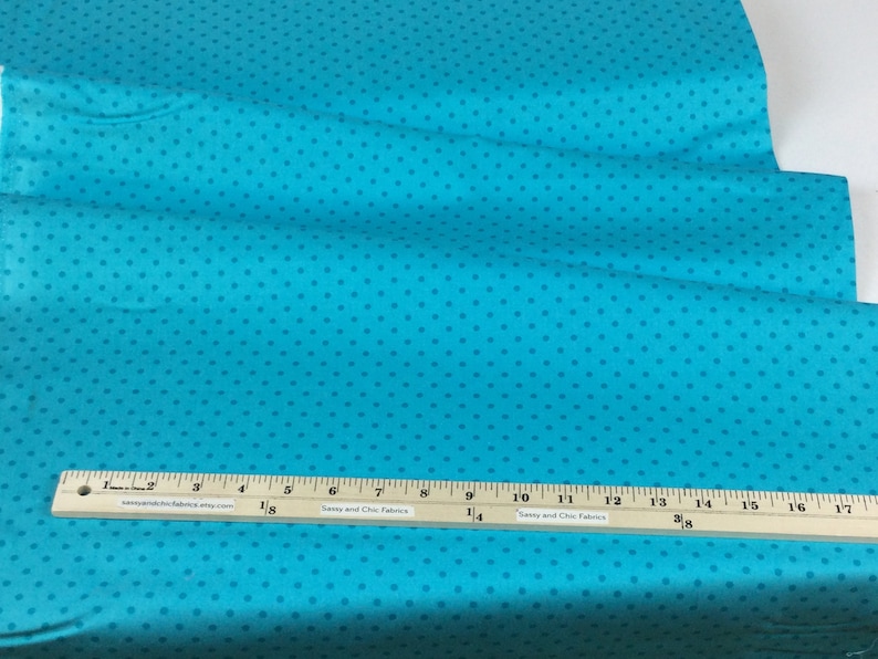 May include: Turquoise fabric with a repeating pattern of small, darker blue dots. A wooden ruler with inch markings is placed across the fabric. The ruler has text that reads "Sassy and Chic Fabrics".