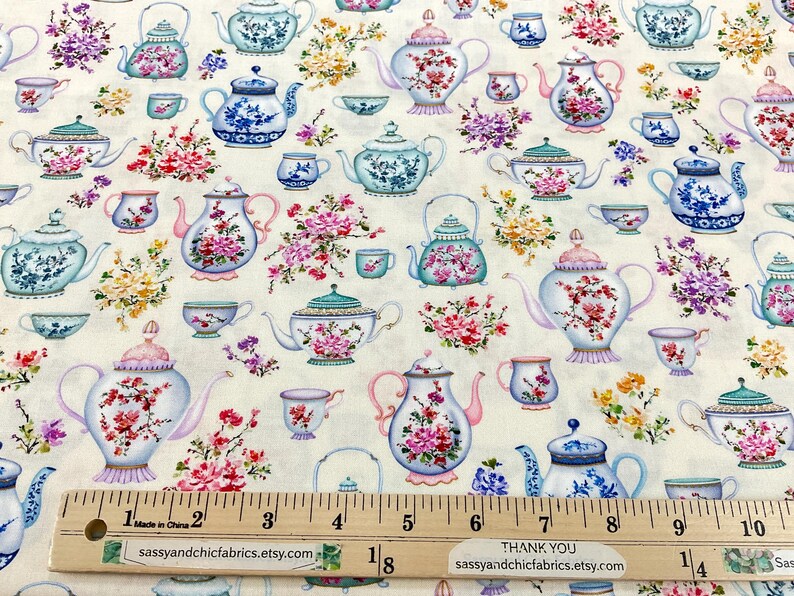 May include: Fabric with a repeating pattern of teapots, teacups, and floral arrangements in shades of blue, pink, and green on a cream background. The fabric includes a ruler with measurements in inches and the text "sassyandchicfabrics.etsy.com".
