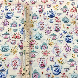 May include: Cream-colored fabric patterned with various teapots and teacups in blue, pink, and green, adorned with floral arrangements. The teapots and teacups are of different shapes and sizes. A wooden ruler is placed vertically on the fabric.