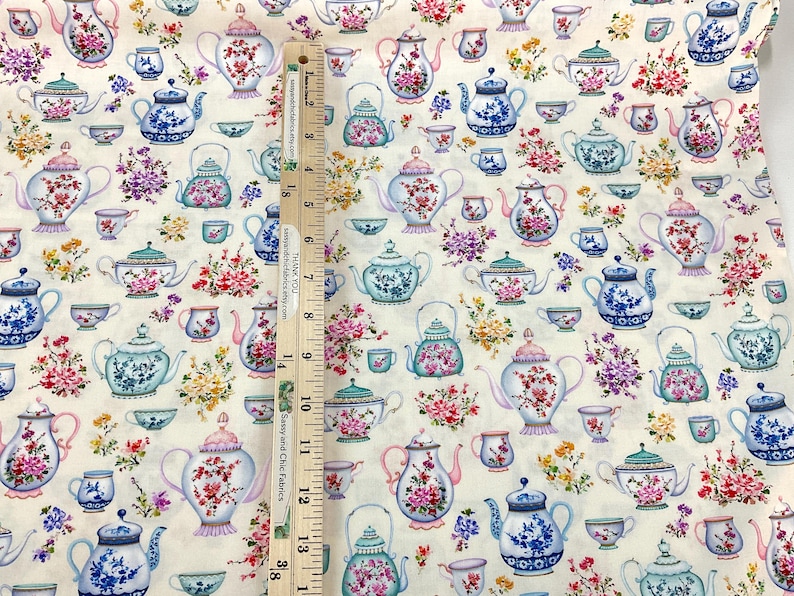 May include: Cream-colored fabric patterned with various teapots and teacups in blue, pink, and green, adorned with floral arrangements. The teapots and teacups are of different shapes and sizes. A wooden ruler is placed vertically on the fabric.