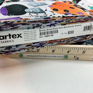 May include: A stack of fabric featuring a Halloween-themed print with gnomes, tombstones, and bats in orange, purple, and black on a gray background. The fabric is labeled "Spooky Gnome Scenic Gray" and measures 10 yards.