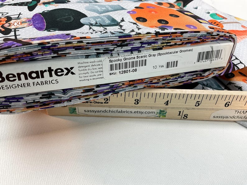 May include: A stack of fabric featuring a Halloween-themed print with gnomes, tombstones, and bats in orange, purple, and black on a gray background. The fabric is labeled "Spooky Gnome Scenic Gray" and measures 10 yards.