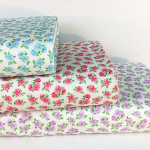 May include: Stacks of floral fabric rolls in blue, pink, and purple rose patterns on a white background. The fabric is tightly rolled, showcasing the detailed floral designs and color variations. Ideal for sewing and crafting projects.