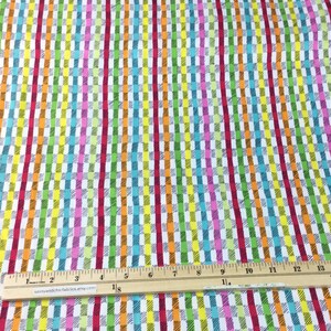 May include: Colorful fabric with a repeating pattern of vertical stripes and rectangular blocks in various colors including red, orange, yellow, green, blue, and pink. A wooden ruler with inch markings is placed on the fabric.