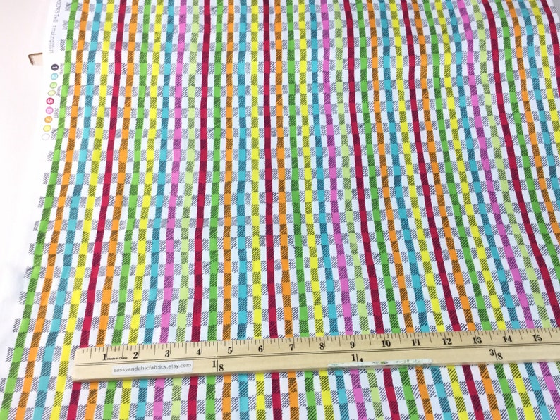 May include: Colorful fabric with a repeating pattern of vertical stripes and rectangular blocks in various colors including red, orange, yellow, green, blue, and pink. A wooden ruler with inch markings is placed on the fabric.