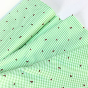 K&ouml;nnte beinhalten: Gr&uuml;n-wei&szlig;es Vichy-Stoff mit einem sich wiederholenden Muster aus Wassermelonenscheiben. Der Stoff ist gefaltet und geschichtet, wodurch das Design zur Geltung kommt. Die Wassermelonenscheiben sind rot mit schwarzen Samen.
