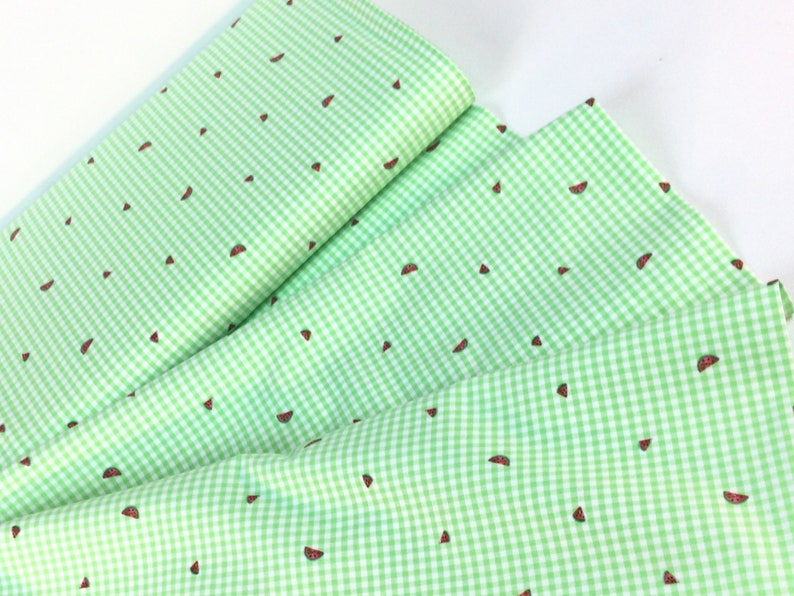 K&ouml;nnte beinhalten: Gr&uuml;n-wei&szlig;es Vichy-Stoff mit einem sich wiederholenden Muster aus Wassermelonenscheiben. Der Stoff ist gefaltet und geschichtet, wodurch das Design zur Geltung kommt. Die Wassermelonenscheiben sind rot mit schwarzen Samen.