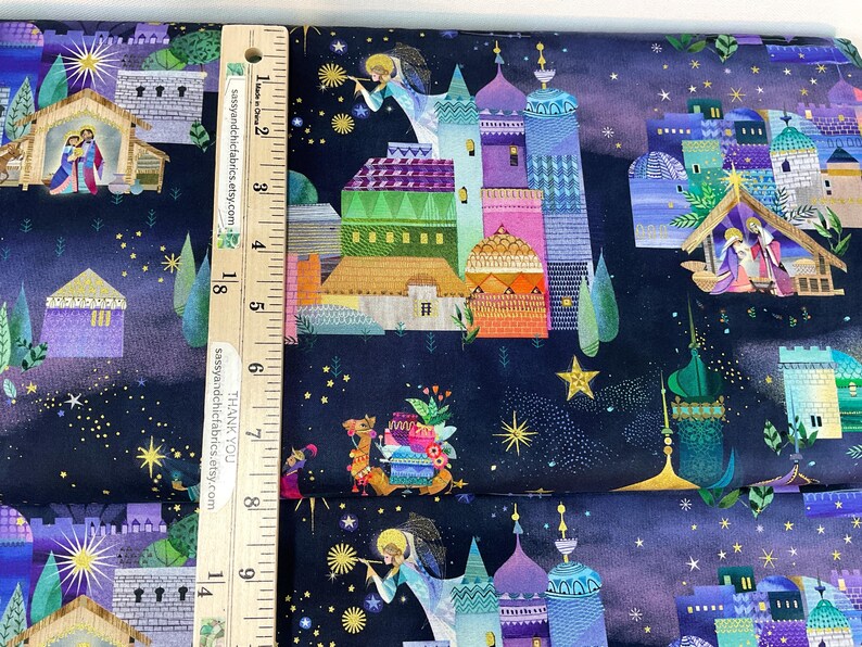 Puede incluir: Tela con un dise&ntilde;o colorido con tem&aacute;tica navide&ntilde;a. La tela presenta ilustraciones de edificios, &aacute;ngeles, estrellas y un pesebre. La paleta de colores incluye azul, morado, verde, naranja y amarillo. Una regla de madera est&aacute; colocada sobre la tela.