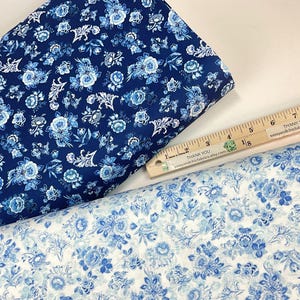May include: Two rolls of floral fabric in shades of blue and white. One roll is a dark navy blue with light blue and white floral patterns. The other roll is a light blue and white floral pattern. A wooden ruler is placed on top of the fabric.