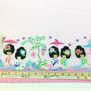 May include: Pink fabric with a repeating pattern of Japanese geisha dolls, pagodas, and floral elements. The design includes parasols, butterflies, and the Japanese character for happiness. A ruler is placed at the bottom.