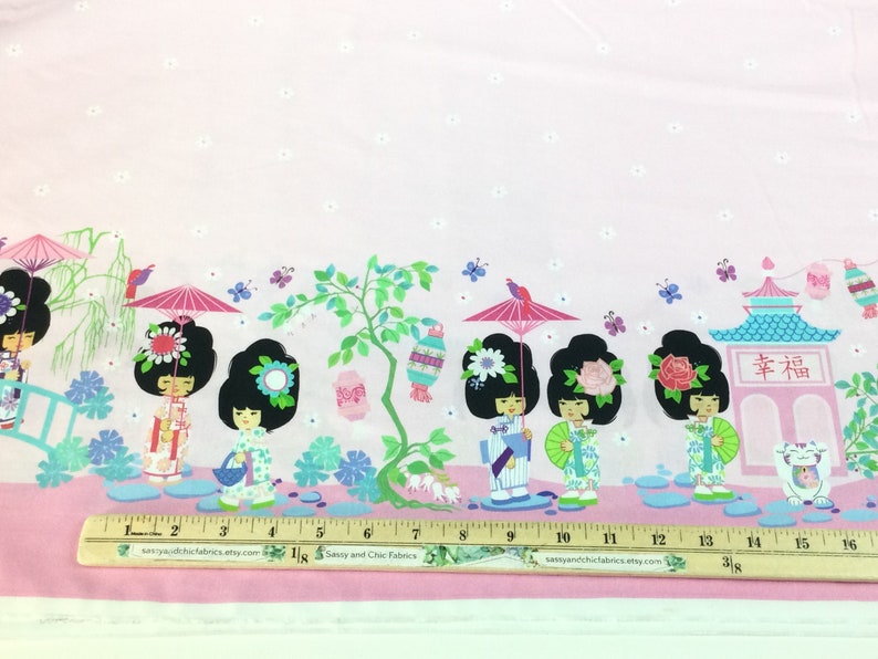 May include: Pink fabric with a repeating pattern of Japanese geisha dolls, pagodas, and floral elements. The design includes parasols, butterflies, and the Japanese character for happiness. A ruler is placed at the bottom.
