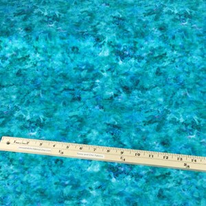 May include: A fabric with a vibrant, abstract design in shades of turquoise, teal, and deep blue. A wooden ruler with inch markings is placed across the fabric. The fabric has a watercolor-like appearance.
