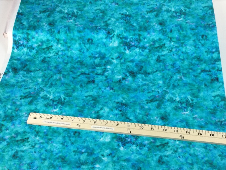 May include: A fabric with a vibrant, abstract design in shades of turquoise, teal, and deep blue. A wooden ruler with inch markings is placed across the fabric. The fabric has a watercolor-like appearance.