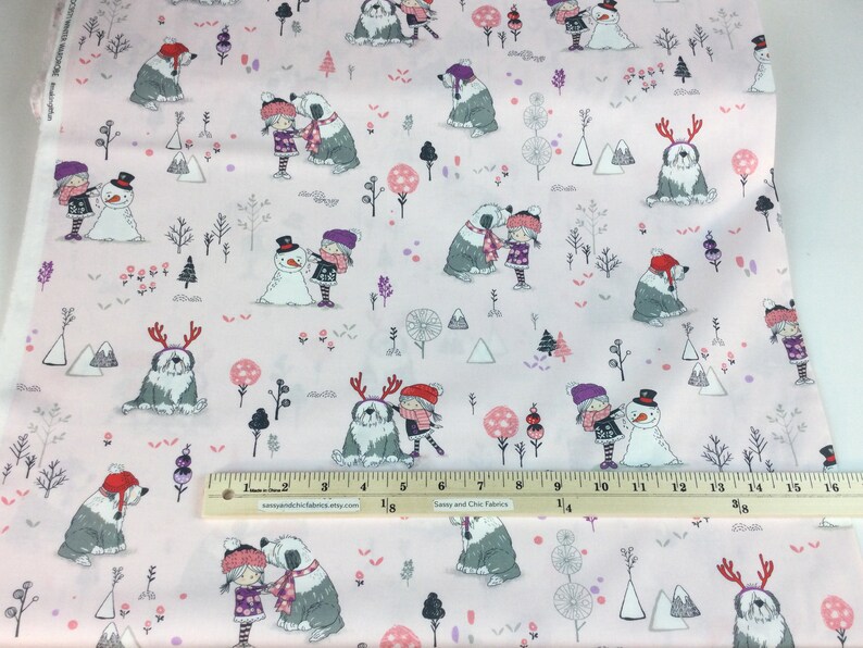 May include: Pink fabric with a winter scene featuring children, snowmen, and Old English Sheepdogs. The design includes trees, mountains, and decorative elements in shades of gray, pink, and purple. A ruler is placed on the fabric.
