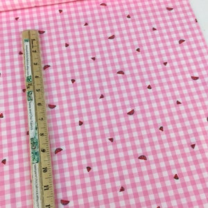 May include: Pink and white gingham fabric with a watermelon slice pattern. The fabric is a light pink color with a classic checkered pattern. Small watermelon slices are scattered across the fabric. A ruler is placed on the fabric.