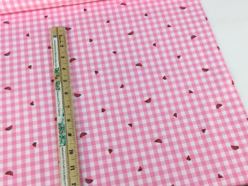 May include: Pink and white gingham fabric with a watermelon slice pattern. The fabric is a light pink color with a classic checkered pattern. Small watermelon slices are scattered across the fabric. A ruler is placed on the fabric.