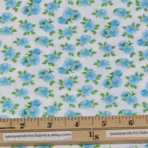 May include: White fabric with a repeating pattern of light blue floral clusters and green leaves. A ruler is placed across the bottom of the fabric, with measurements in inches.