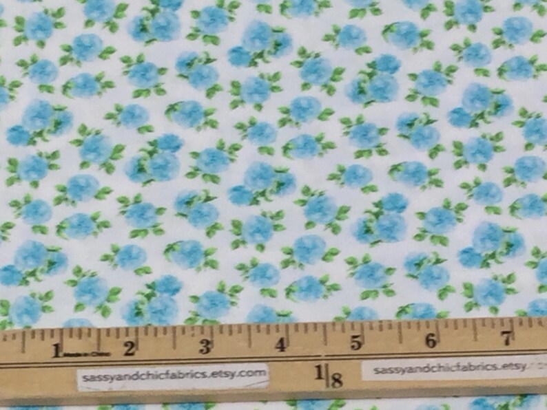 May include: White fabric with a repeating pattern of light blue floral clusters and green leaves. A ruler is placed across the bottom of the fabric, with measurements in inches.
