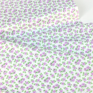 May include: White fabric with a repeating pattern of small, light purple roses and green leaves. The floral design is densely packed across the fabric's surface, creating a delicate and feminine aesthetic. Ideal for sewing projects.