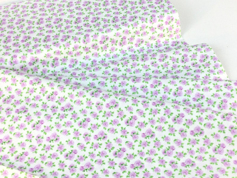 May include: White fabric with a repeating pattern of small, light purple roses and green leaves. The floral design is densely packed across the fabric's surface, creating a delicate and feminine aesthetic. Ideal for sewing projects.