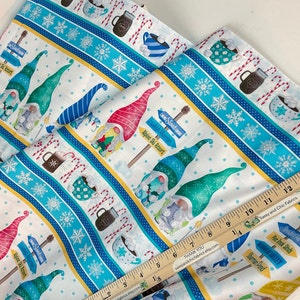 May include: White fabric with a winter-themed pattern featuring gnomes, snowflakes, hot cocoa, and candy canes. The design includes directional signs for an Alpine Ski Resort and other locations. A ruler is included for scale.