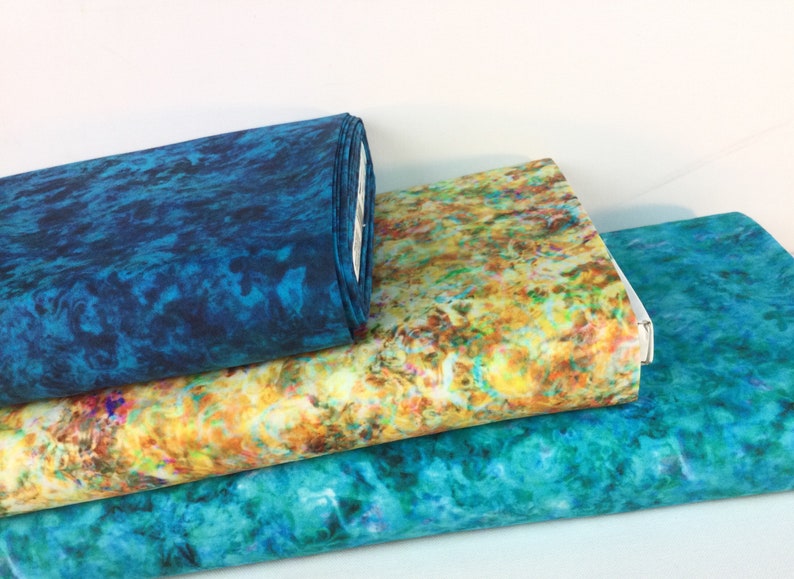May include: Three rolls of fabric with vibrant, abstract patterns. One roll is a deep teal, while the others display a mix of yellow, orange, and teal hues. The fabrics appear to have a marbled or watercolor effect.