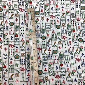 May include: Mahjong tile fabric with a cream background. The fabric features a repeating pattern of Mahjong tiles in various colors, including red, green, and black. A wooden ruler with the text "THANK YOU" is placed on the fabric.
