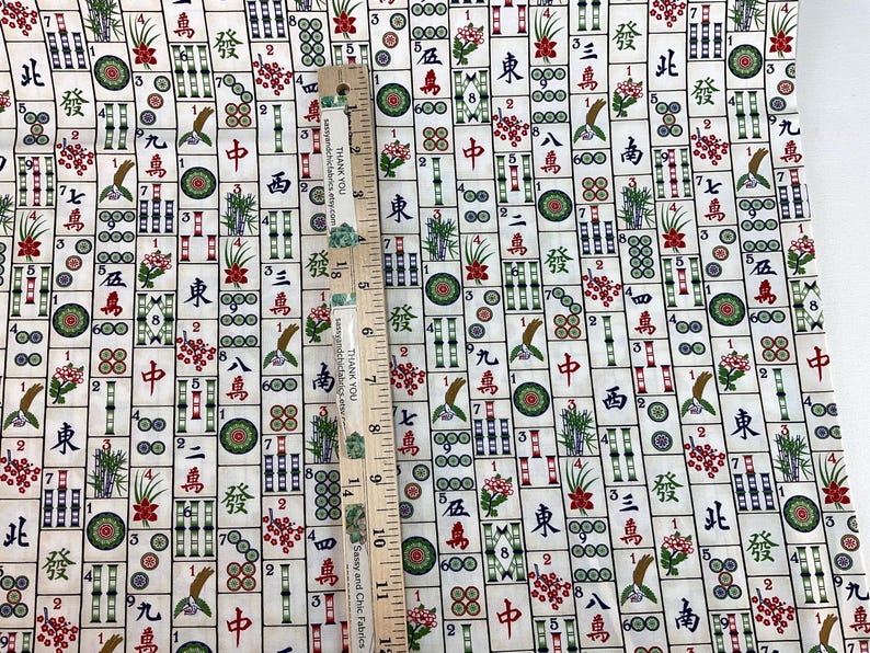 May include: Mahjong tile fabric with a cream background. The fabric features a repeating pattern of Mahjong tiles in various colors, including red, green, and black. A wooden ruler with the text "THANK YOU" is placed on the fabric.
