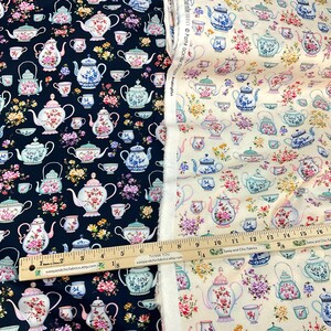 May include: Two fabric samples, one navy blue and one cream, feature a repeating pattern of teapots, teacups, and floral arrangements. The teapots are in various colors, including pink, blue, and green. A wooden ruler with inch measurements is visible.