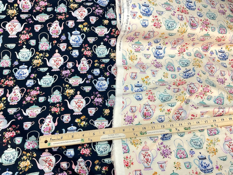 May include: Two fabric samples, one navy blue and one cream, feature a repeating pattern of teapots, teacups, and floral arrangements. The teapots are in various colors, including pink, blue, and green. A wooden ruler with inch measurements is visible.