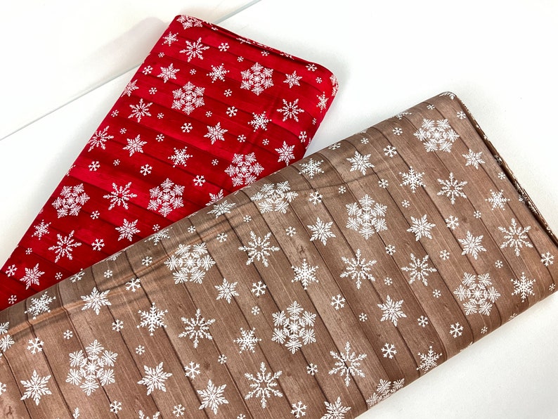 Pu&ograve; includere: Due rotoli di tessuto a tema invernale. Uno &egrave; rosso con fiocchi di neve bianchi e un motivo a venature di legno. L'altro &egrave; marrone con fiocchi di neve bianchi e un motivo a venature di legno. Il tessuto &egrave; piegato.