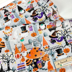 May include: Halloween-themed fabric featuring gnomes, pumpkins, witch hats, and tombstones on a light gray background. The fabric includes orange, black, purple, and white elements, with a ruler in the bottom right corner.