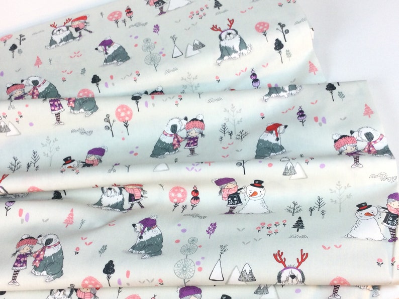 May include: Light blue fabric with a winter scene print. The design features children, dogs, snowmen, trees, and mountains in shades of gray, pink, and purple. The fabric is folded, showing the pattern's repeat.