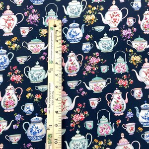 May include: Fabric with a navy blue background featuring a repeating pattern of teapots and teacups in various designs. The teapots and cups are decorated with floral patterns in pink, white, and blue. A ruler is placed vertically on the fabric.