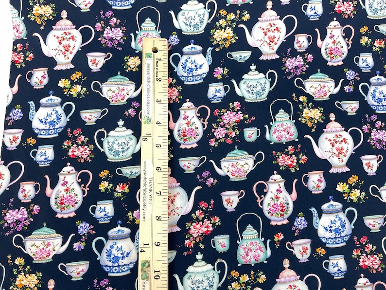 May include: Fabric with a navy blue background featuring a repeating pattern of teapots and teacups in various designs. The teapots and cups are decorated with floral patterns in pink, white, and blue. A ruler is placed vertically on the fabric.