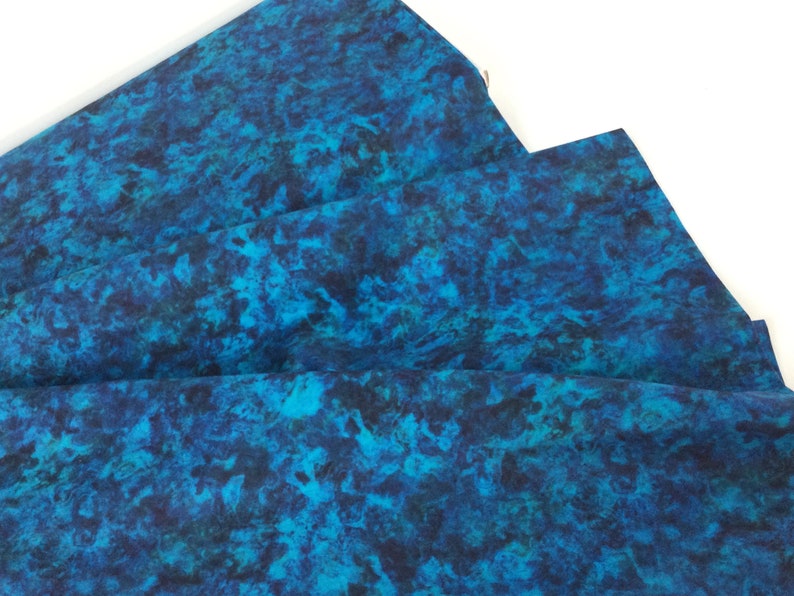 May include: Folded fabric with a vibrant, abstract pattern in shades of teal, turquoise, and deep blue. The design resembles a watercolor effect or a marbled appearance. The fabric is likely used for sewing or crafting projects.
