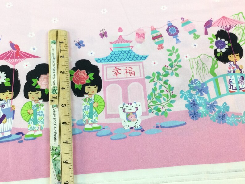 May include: Pink fabric with a Japanese-themed print. The design features cartoon geishas, pagodas, lucky cats, and floral elements. A wooden ruler is placed on the fabric, showing measurements in inches.