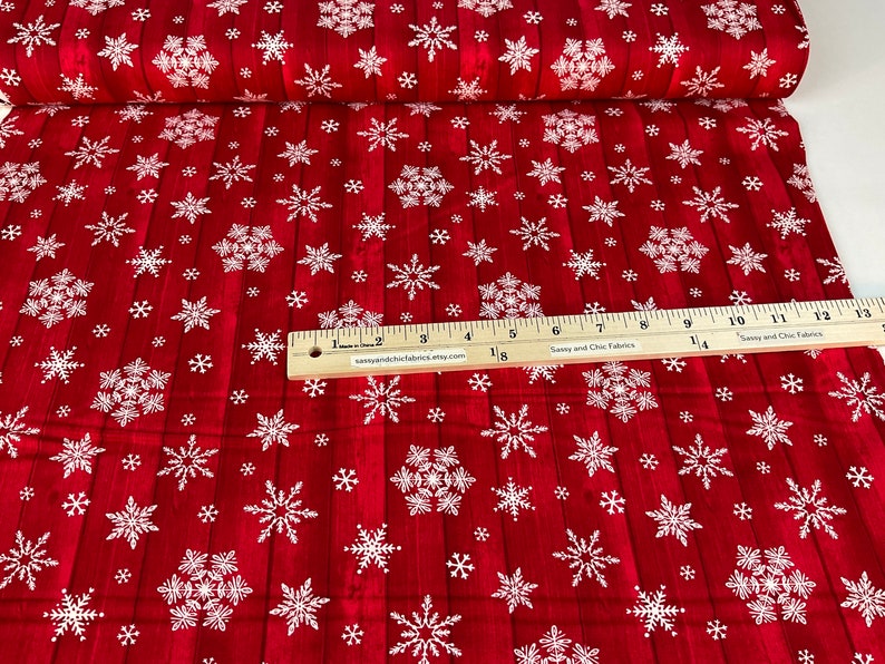 Pu&ograve; includere: Tessuto rosso con motivo a fiocchi di neve bianchi. Il tessuto presenta un effetto legno e un righello &egrave; posizionato sopra. Il testo "Sassy and Chic Fabrics" &egrave; visibile sul righello. Adatto per lavori creativi natalizi.