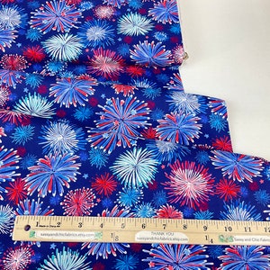 May include: A bolt of fabric with a navy blue background, featuring a repeating pattern of red, white, and blue fireworks. A wooden ruler with inch measurements is placed on the fabric. The text "Thank You" and "Sassy and Chic Fabrics" are visible.