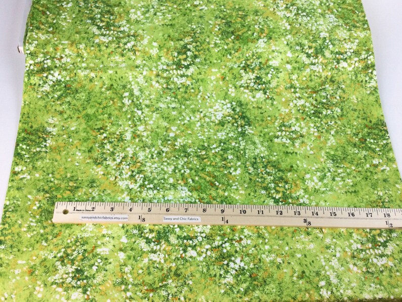 May include: Green fabric with a meadow-like pattern of white and yellow flowers. A wooden ruler with inch markings is placed on the fabric. The text "Sassy and Chic Fabrics" is visible on the ruler.