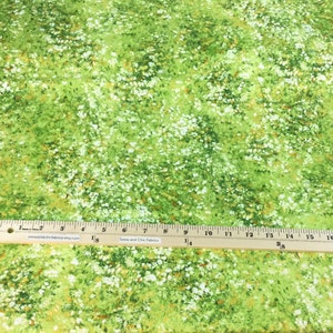 May include: Green fabric with a floral pattern featuring white and yellow accents. A wooden ruler with inch measurements is placed across the fabric. The fabric has a meadow-like appearance.