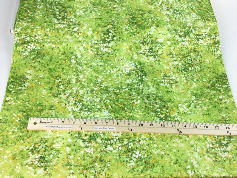 May include: Green fabric with a floral pattern featuring white and yellow accents. A wooden ruler with inch measurements is placed across the fabric. The fabric has a meadow-like appearance.