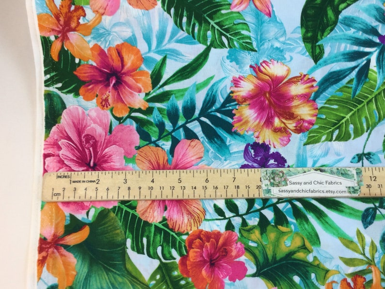 May include: Fabric with a tropical floral pattern featuring large hibiscus flowers in shades of pink, orange, and yellow, set against a light blue background with green leaves. A ruler shows measurements in inches.