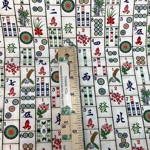 May include: Fabric with a repeating pattern of Mahjong tiles in cream, red, green, and black. The design includes Chinese characters, bamboo, flowers, and circles. A wooden ruler is placed on the fabric.