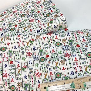 May include: White fabric with a repeating pattern of Mahjong tiles in red, green, and blue. The fabric includes Chinese characters and floral designs. A wooden ruler with inch markings is in the foreground.