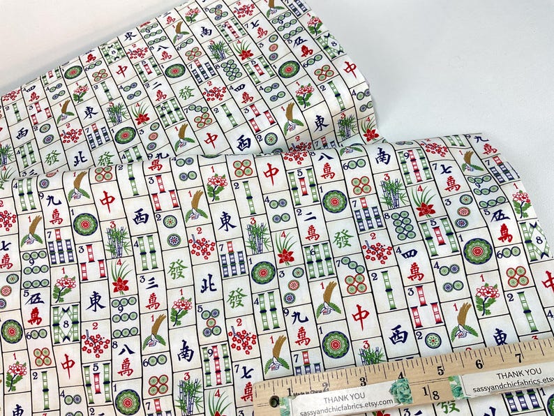 May include: White fabric with a repeating pattern of Mahjong tiles in red, green, and blue. The fabric includes Chinese characters and floral designs. A wooden ruler with inch markings is in the foreground.